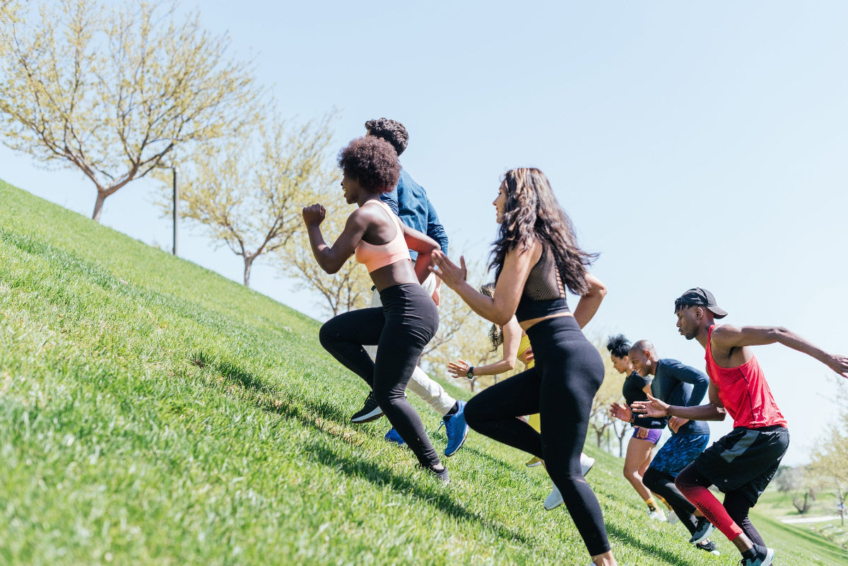 Diverse young adult running group runs up a green park hill on a hot sunny summer day with smiles on their faces.