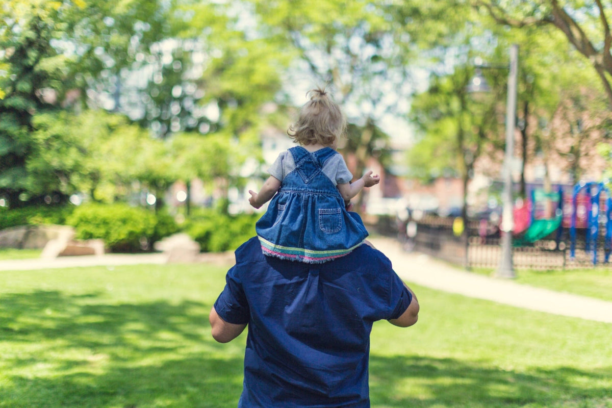 Young toddler daughter sits on her father's shoulders at green New York park playground in the summer sun