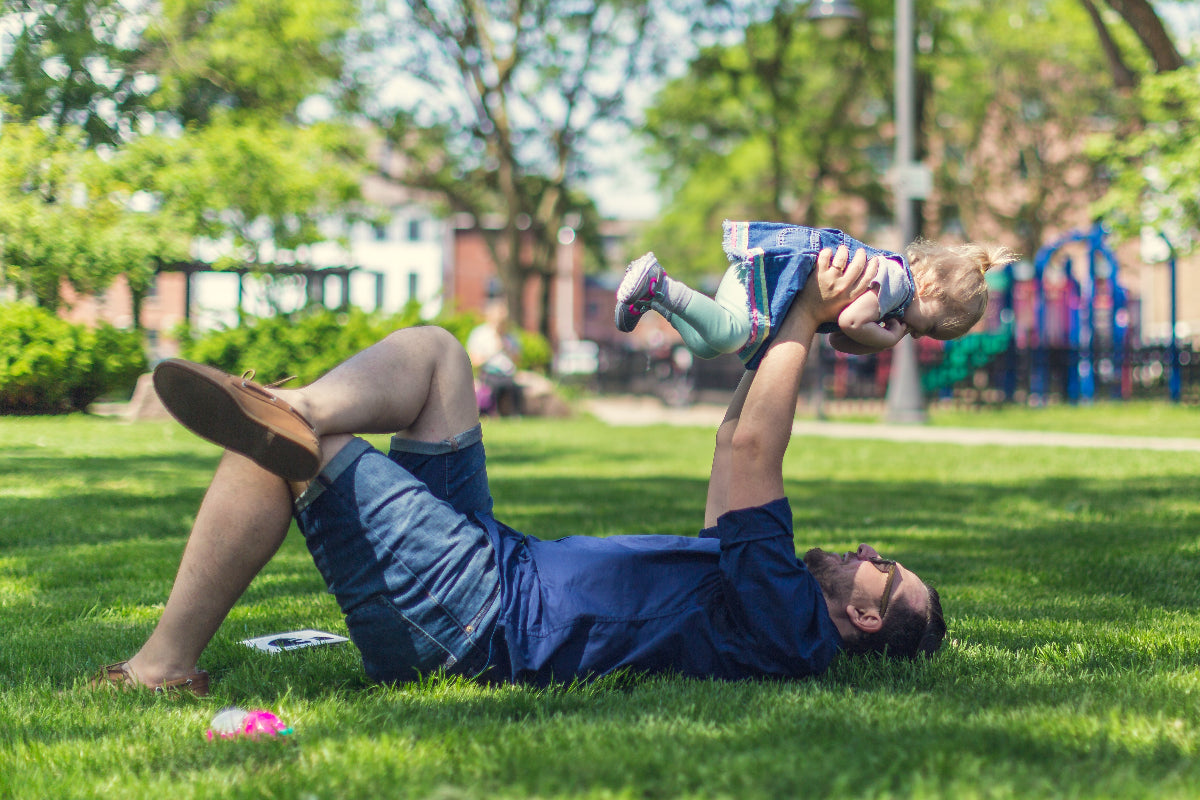 dad lying down and holding baby toddler daughter in the air at the park in the grass next to the playground