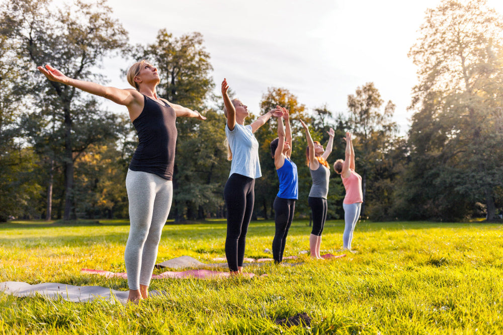 Group of five women doing yoga and stretches outdoor on grass in forest park.