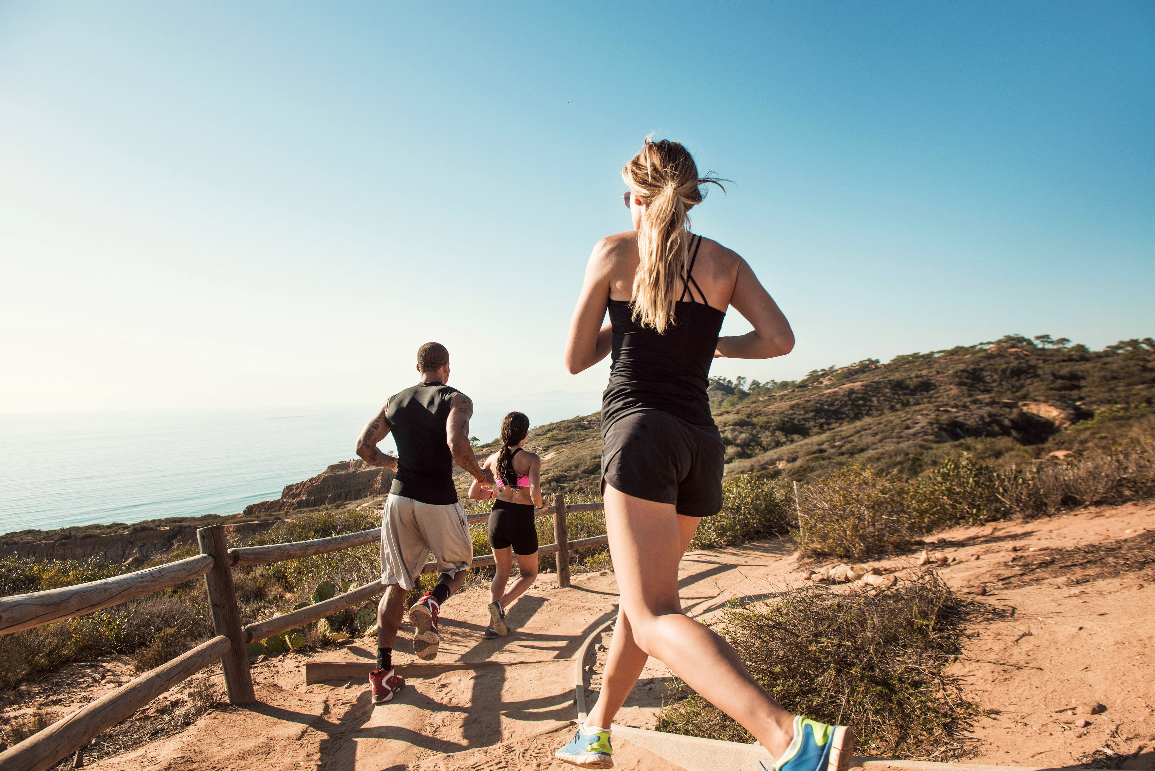 A mixed-race group of friends run down a California desert trail in the hot sun on a summer's day with a view of the hills and ocean.