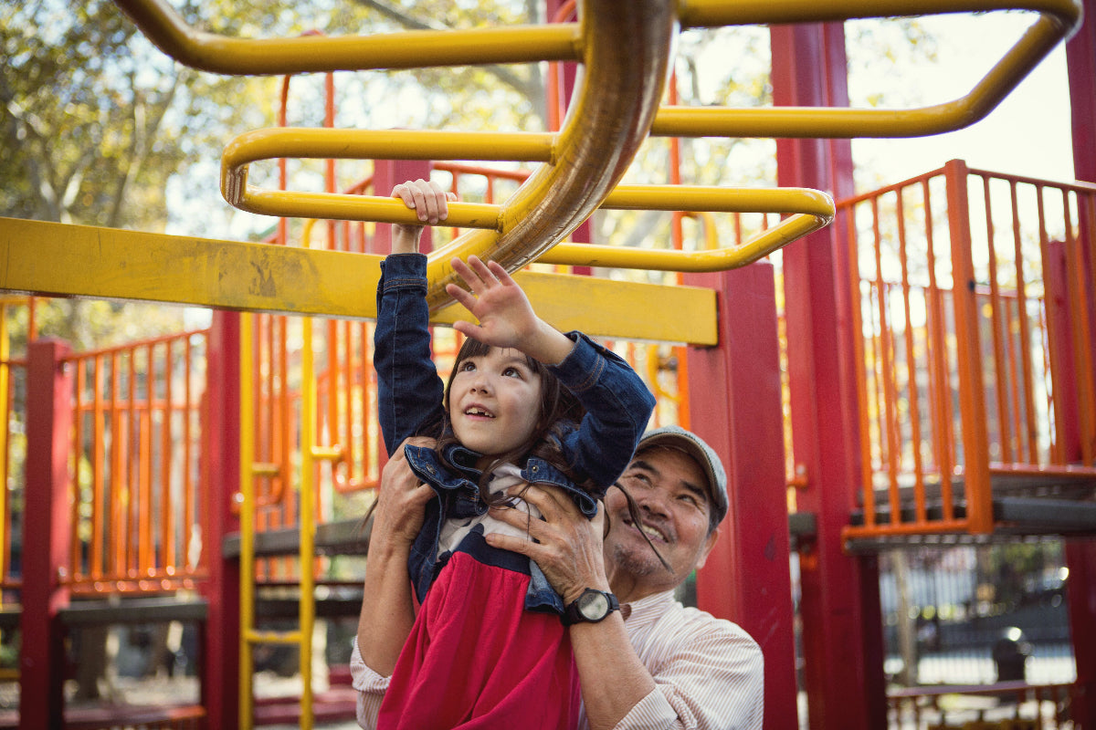 Grandpa holds granddaughter up to climb yellow monkey bars at outdoor children's playground
