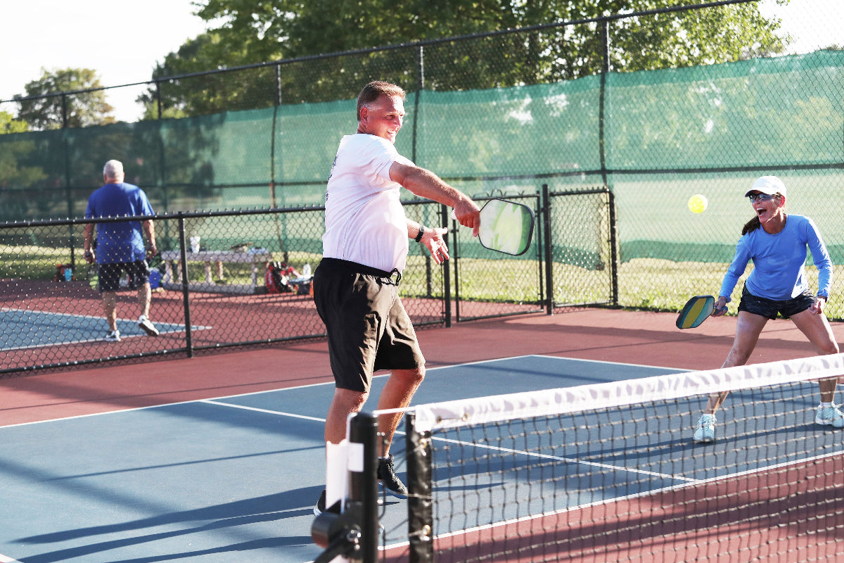 An older senior man with flushed rosy cheeks smiles while playing outdoor pickleball with friends at the local pickleball tennis courts in the park
