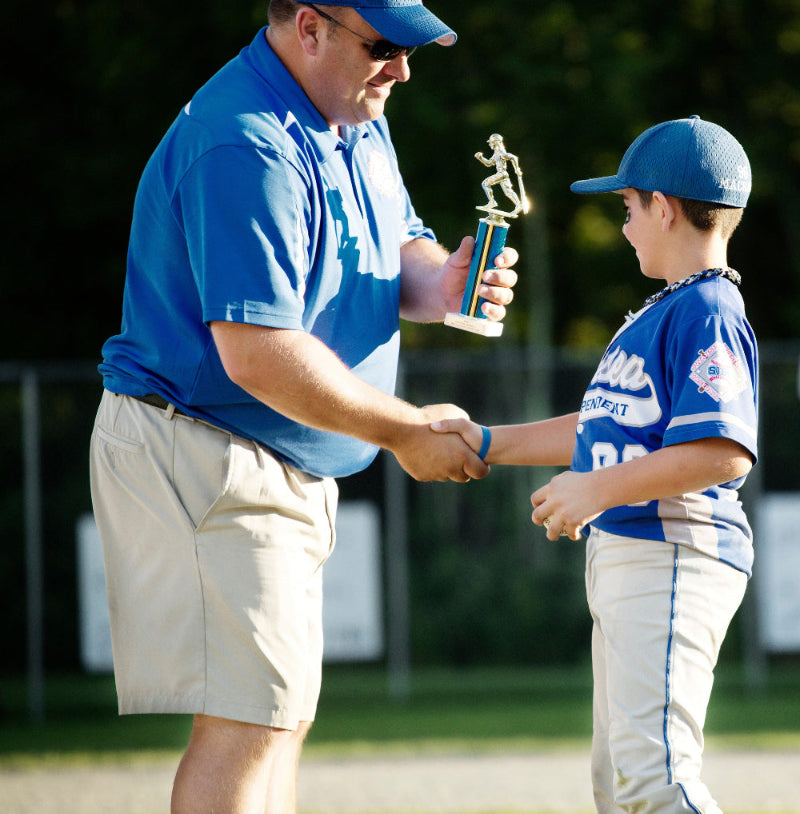 Coach and young boy in blue junior baseball uniforms shaking hands with a trophy on a sports field.
