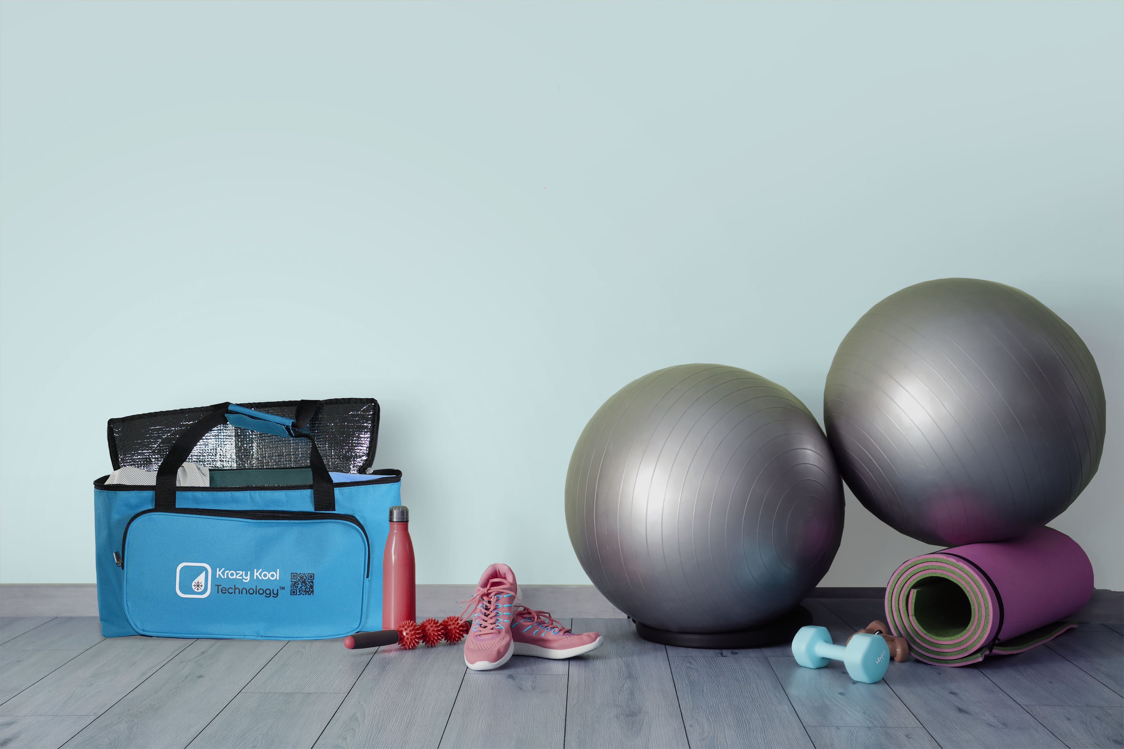 Gym equipment including a blue bag, water bottle, sneakers, exercise balls, and a yoga mat on a wooden floor.