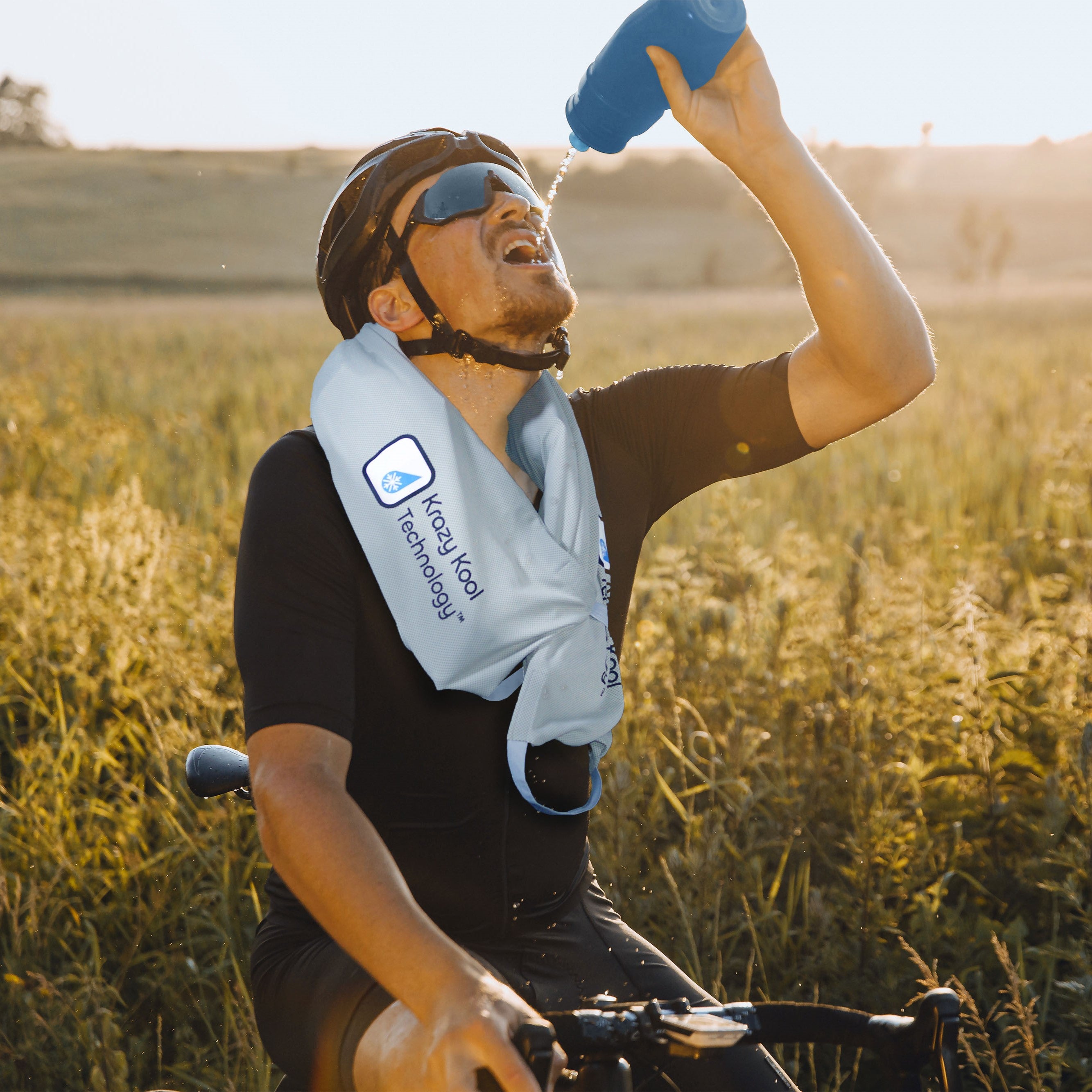 Long-distance thirsty and sweating male cyclist drinking from blue water bottle wearing Krazy Kool Technology System on bike ride through highway roads and farm fields.